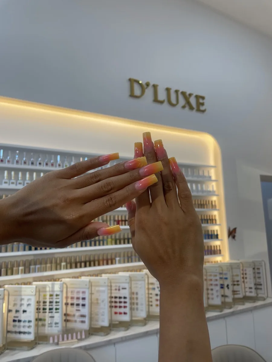Two hands with orange and yellow ombre nails raised in front of the salon's backlit wall of polish bottles