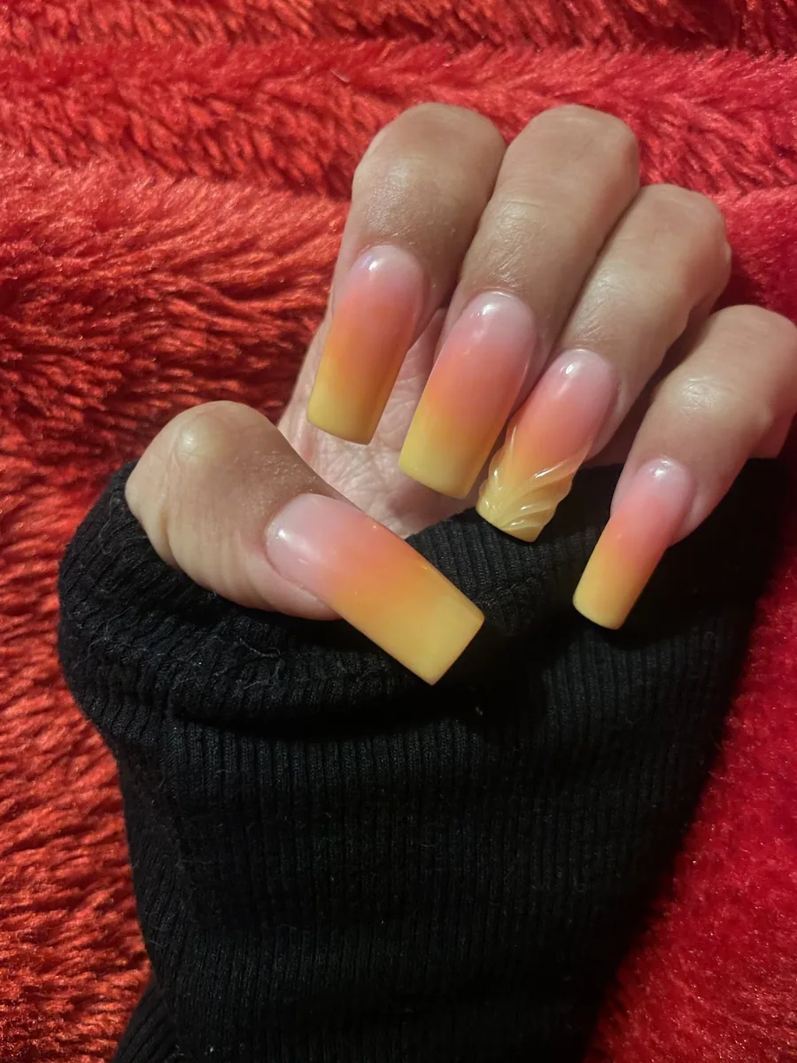 Close-up of a hand in a black sleeve on red fabric with long square peach-to-yellow ombre nails